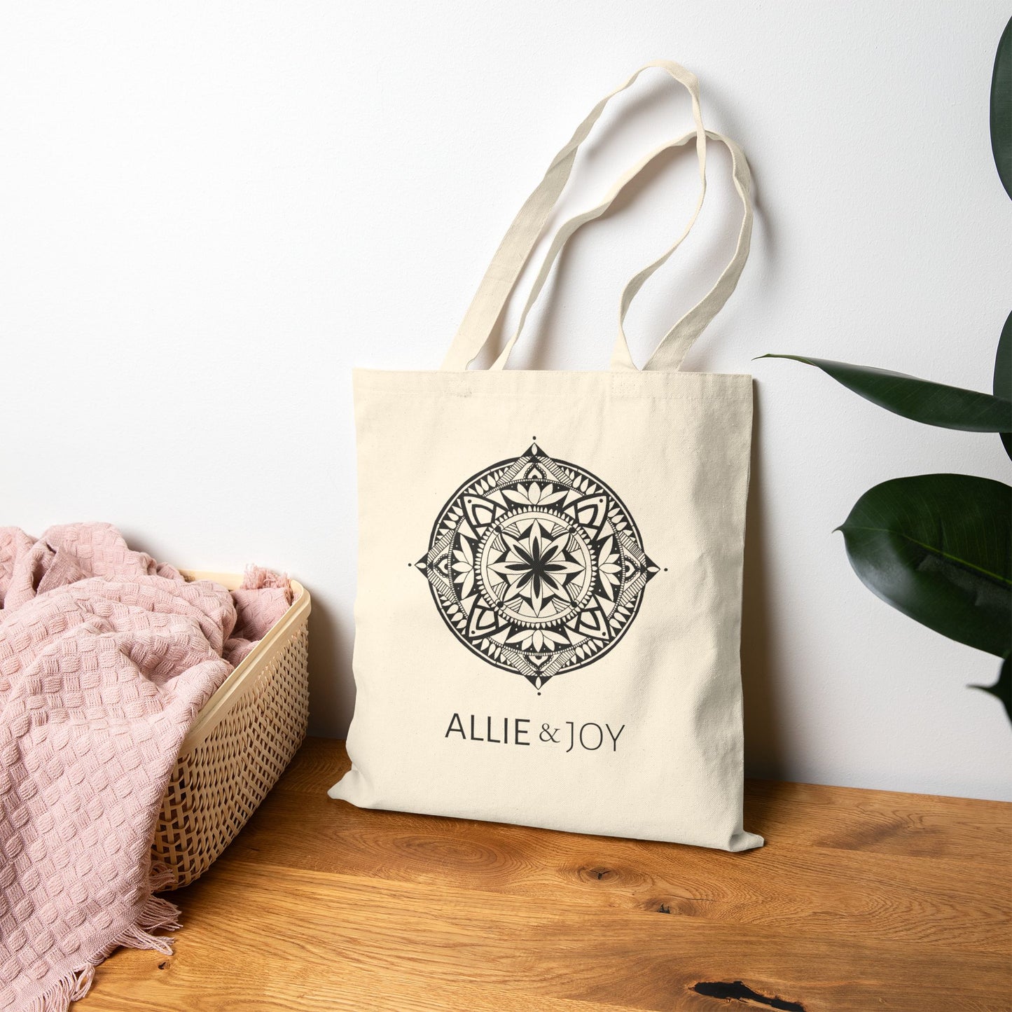 Beige tote bag with mandala pattern and 'Allie & Joy' logo below on a wooden surface with a white wall and plant in the background