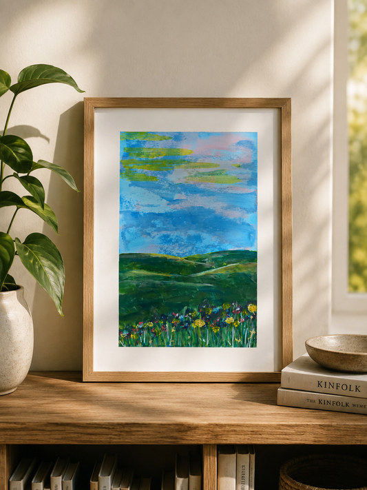 A framed landscape art print featuring green rolling hills and a blue sky with clouds, styled on a wooden shelf next to a potted plant and books.
